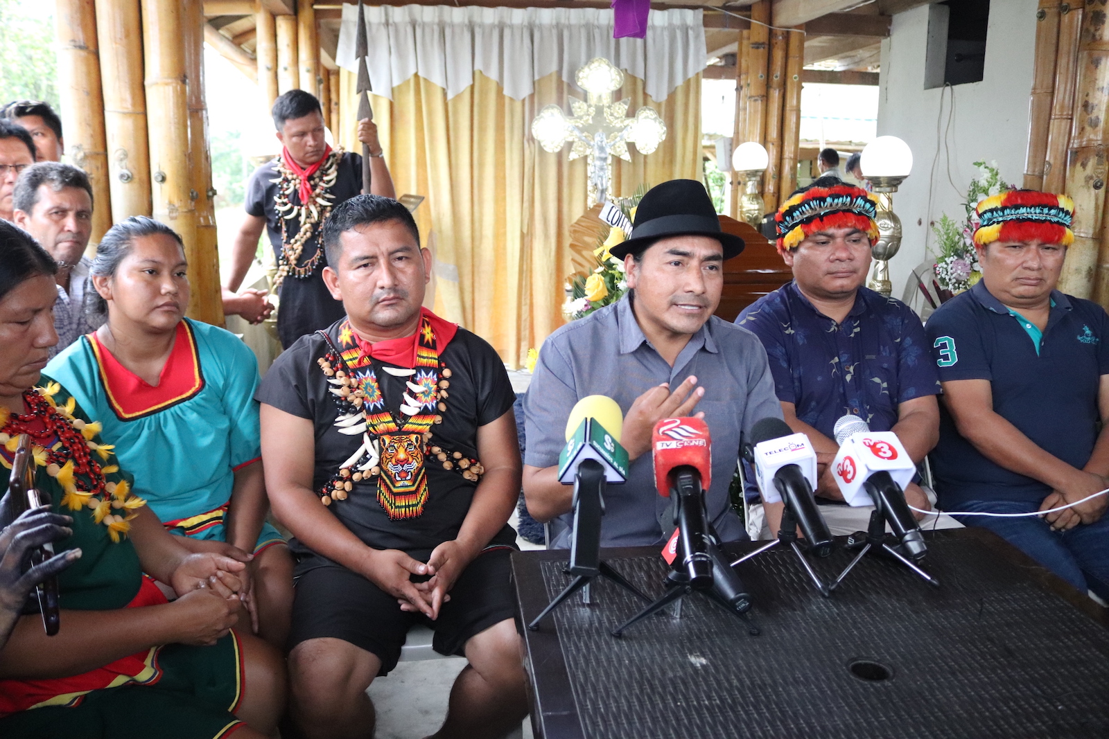 A group of people gather looking somber. A man in a black hat speaks toward a table covered in several microphones. Next to him, a man in a black shirt and elaborate beaded necklaces listens.
