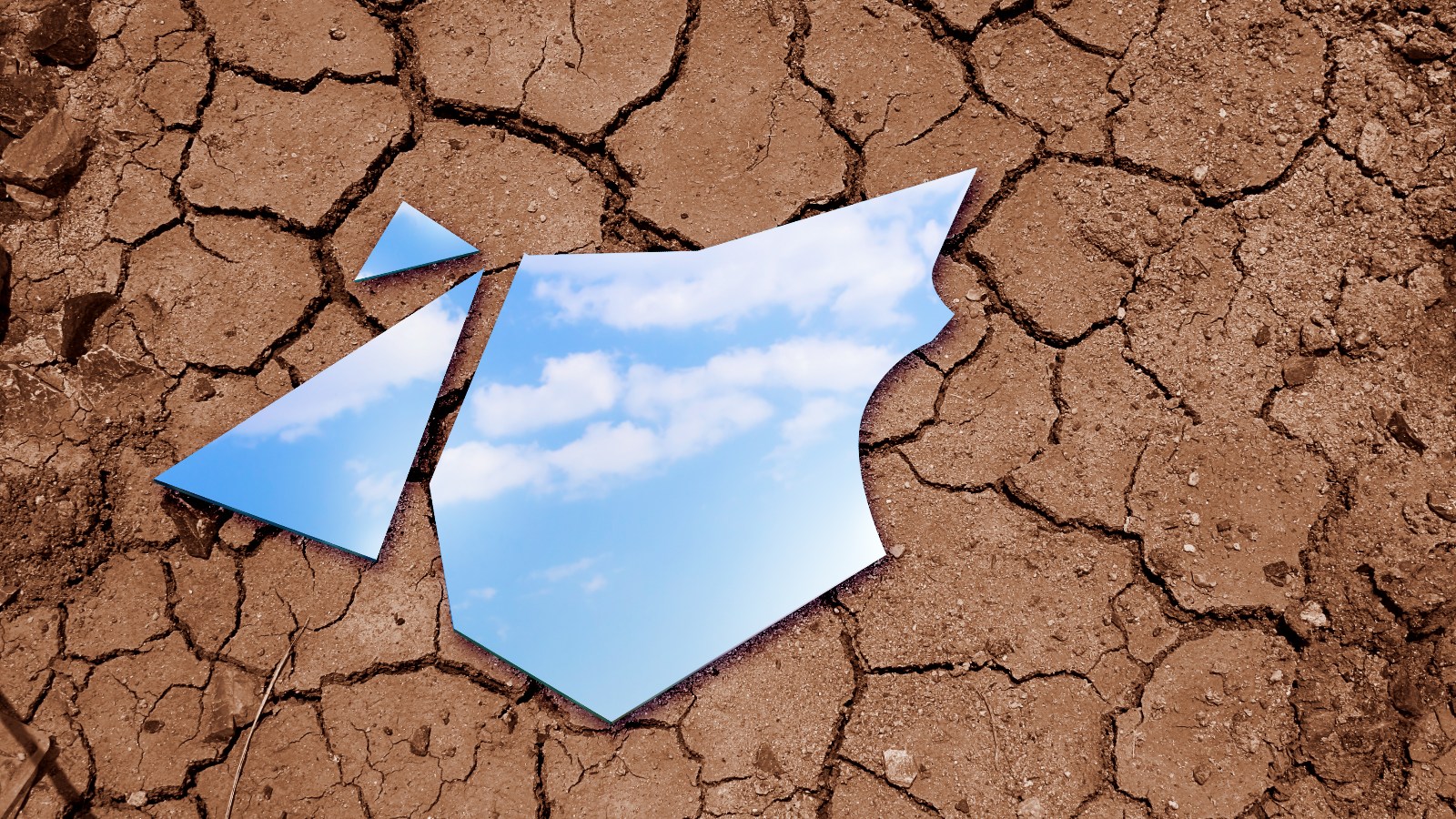 Photo of a shattered mirror placed on dry, cracked soil reflecting a blue sky