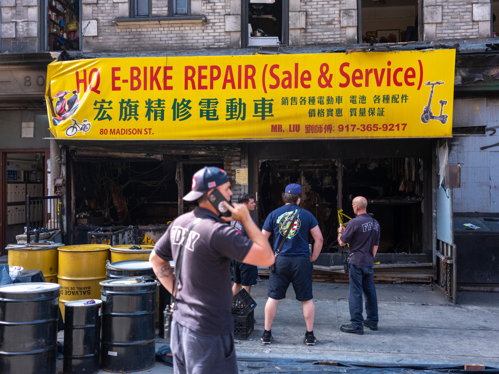 A man wearing an FDNY shirt holds a cell phone to his ear in front of the burned-out shell of a shop under the sign "HQ E-BIKE REPAIR (Sale & Service)"