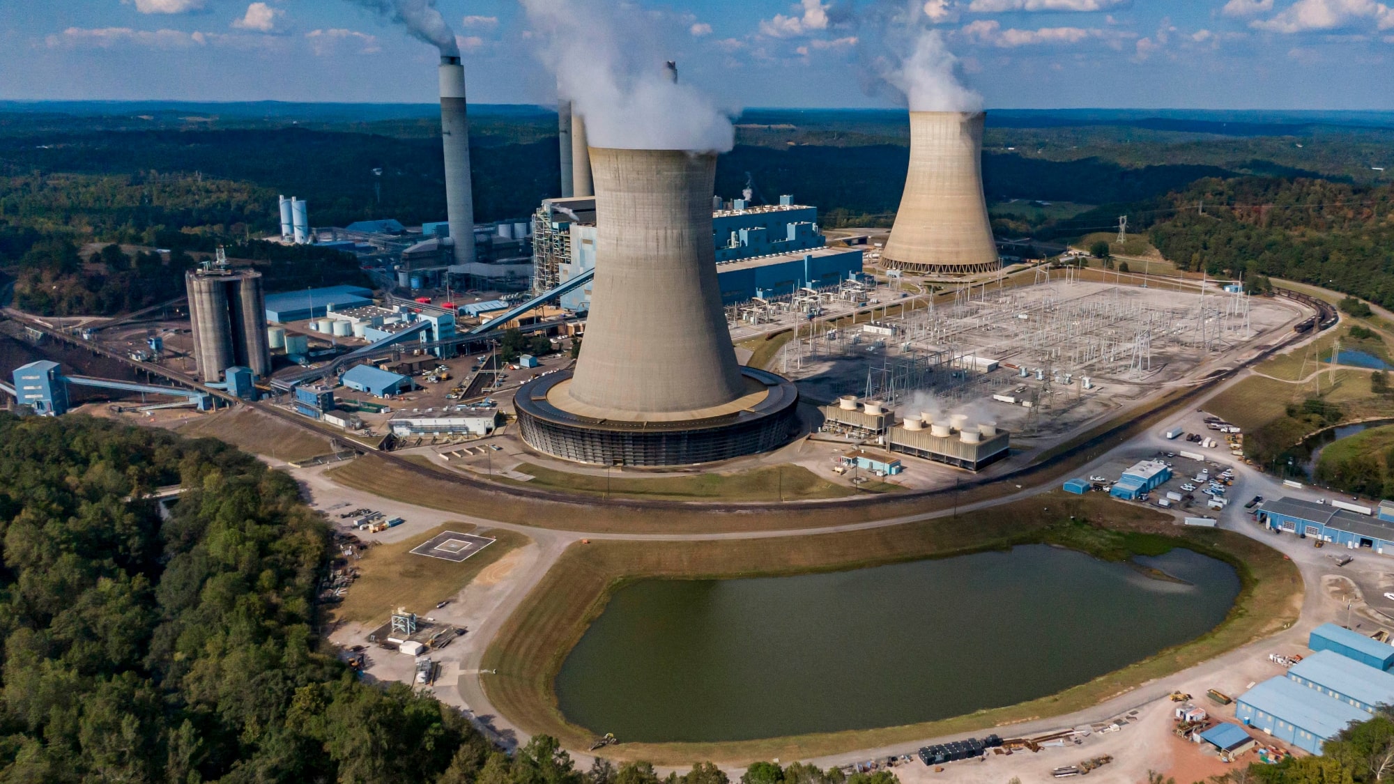 Smoke billows from a large power plant with two huge cooling towers set next to a pond.