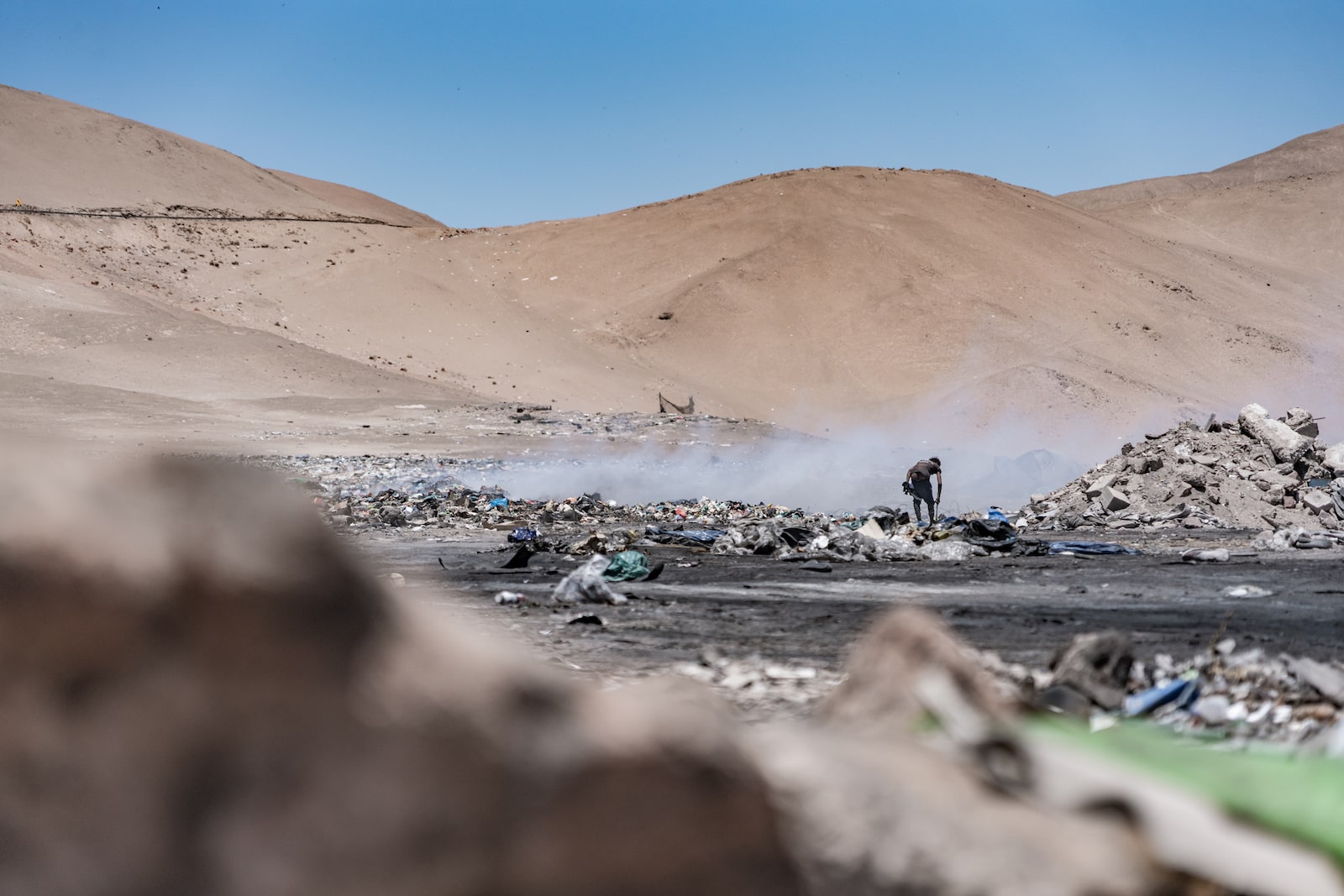 A man stands near large piles of burning clothing in the desert