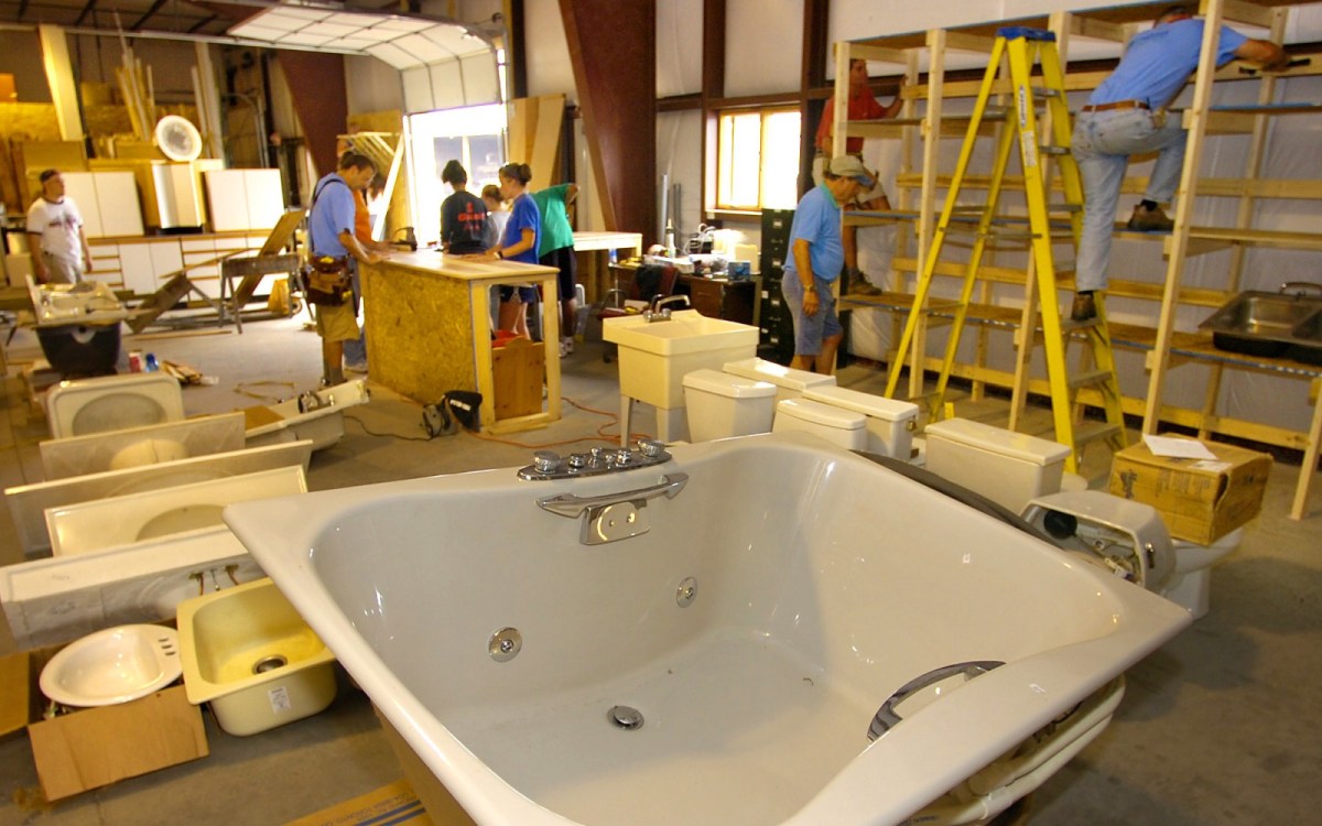 A large bathtub sits in the foreground of a warehouse-style space, along with other plumbing fixtures, with people working to line the corridors.