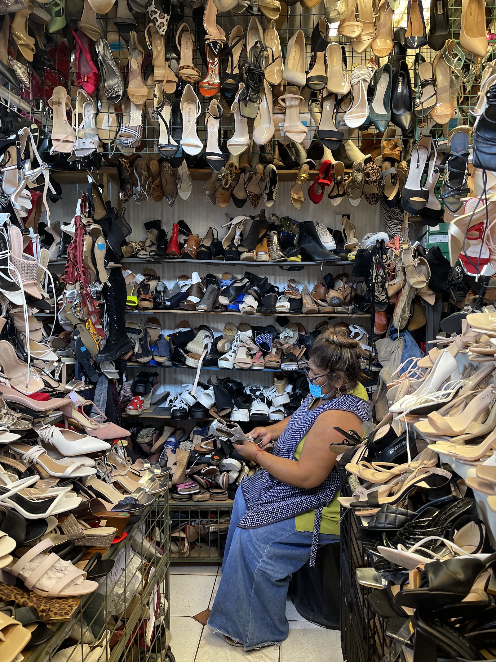 a woman looks toward a wall of used shoes lining the inside of a kiosk in an outdoor market