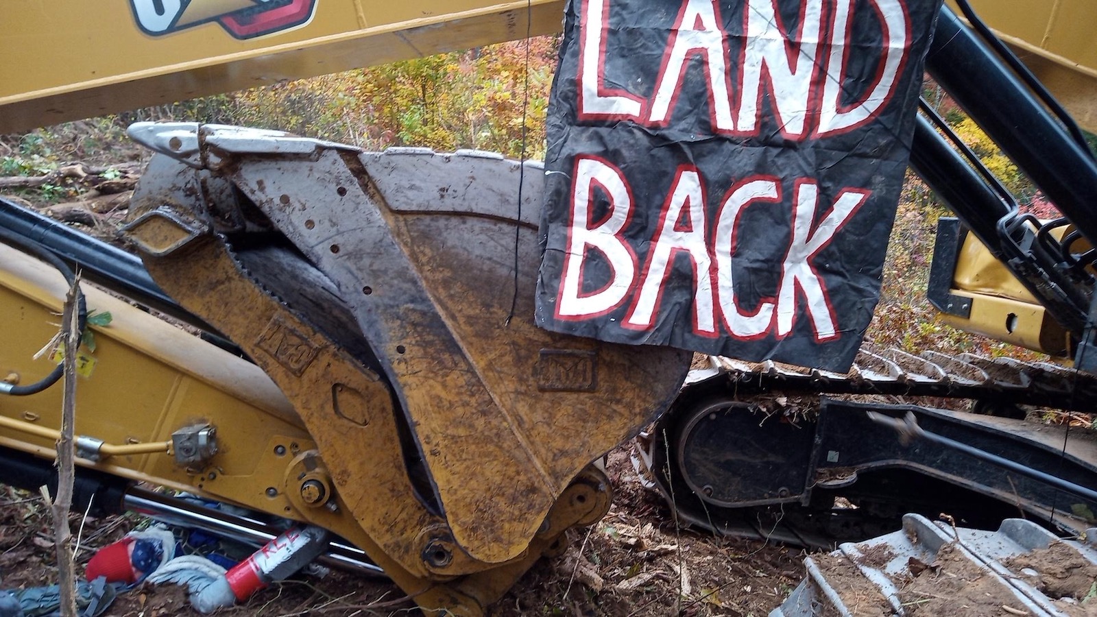 A protestor is chained to a piece of heavy construction equipment beneath a banner reading "Land Back."