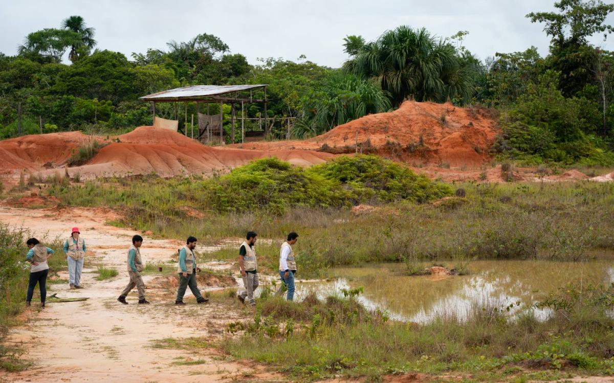 A group of men in beige vests walks down an exposed red dirt road toward a small pond, with tropical foliage in the background.