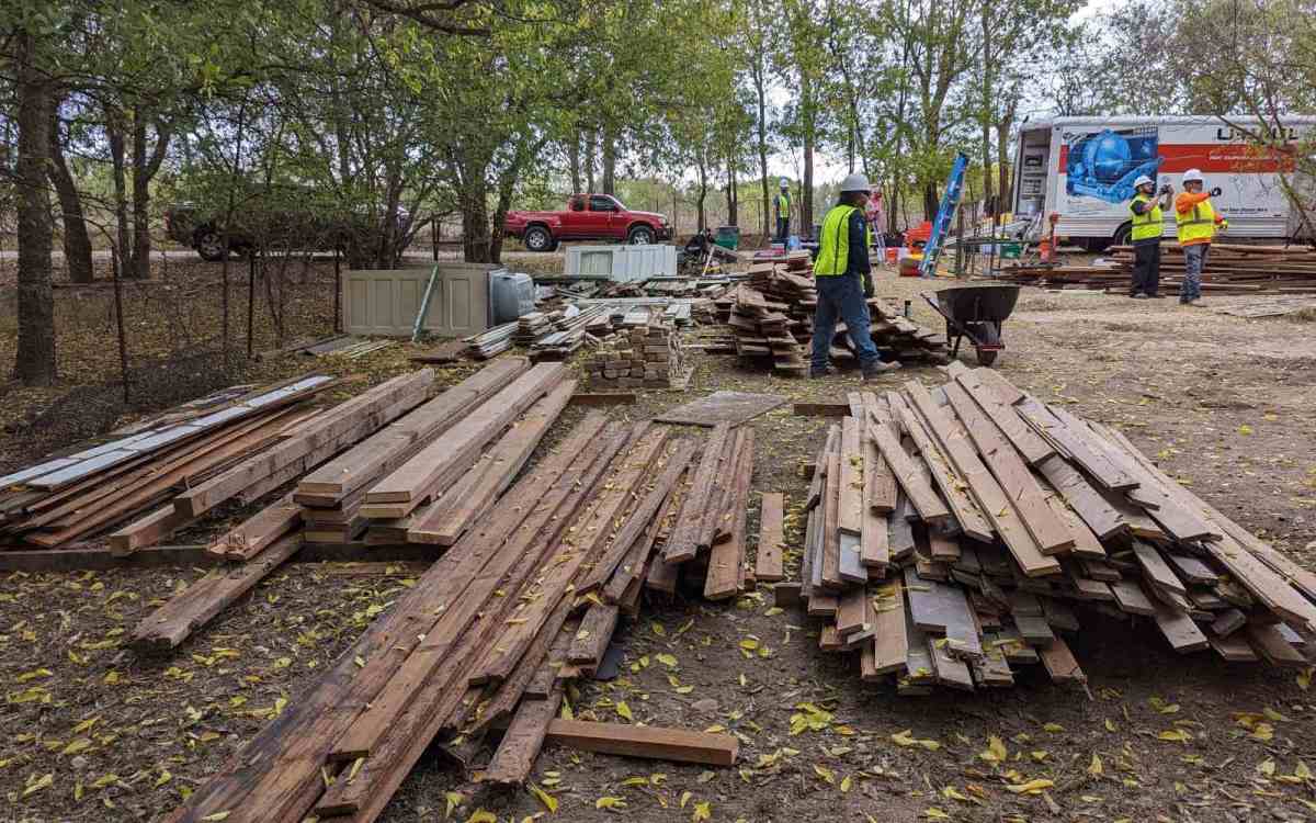 Piles of wooden planks sit in the foreground of a treed area. People in hard hats and reflective vets walk around the piles.
