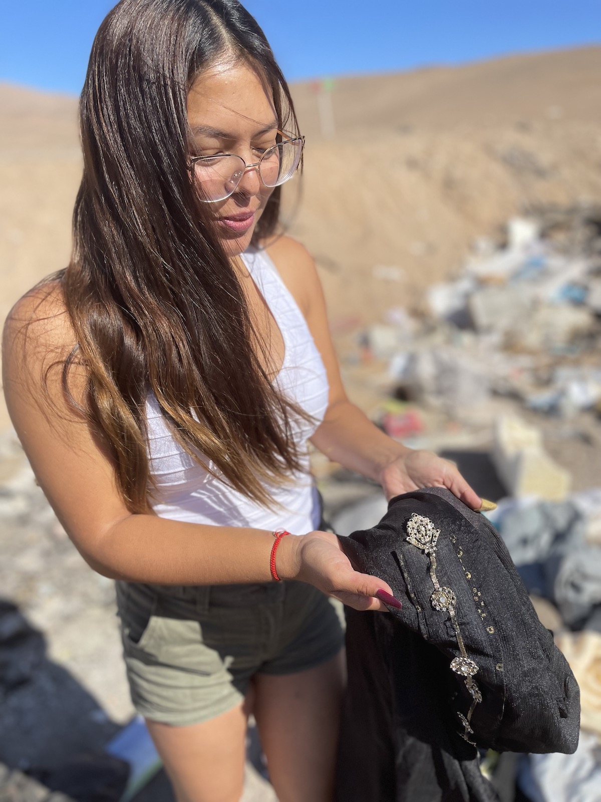 a young woman holds a black piece of clothing with embroidery near a large pile of textile trash in the desert