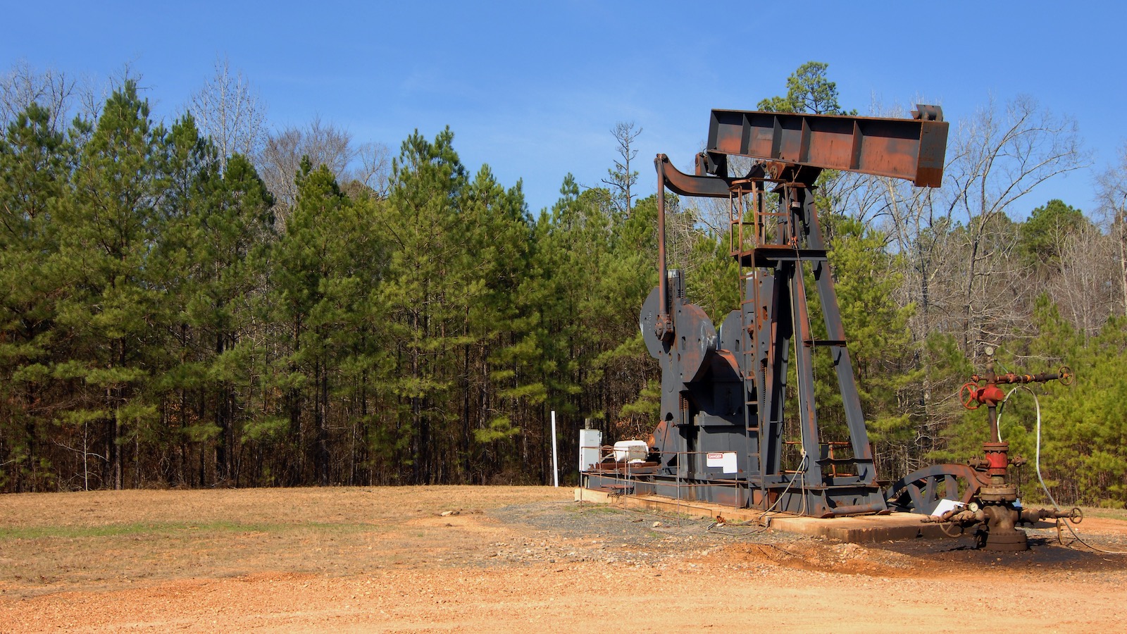 An oil well sits idle surrounded by pine trees and blue sky in rural Arkansas.