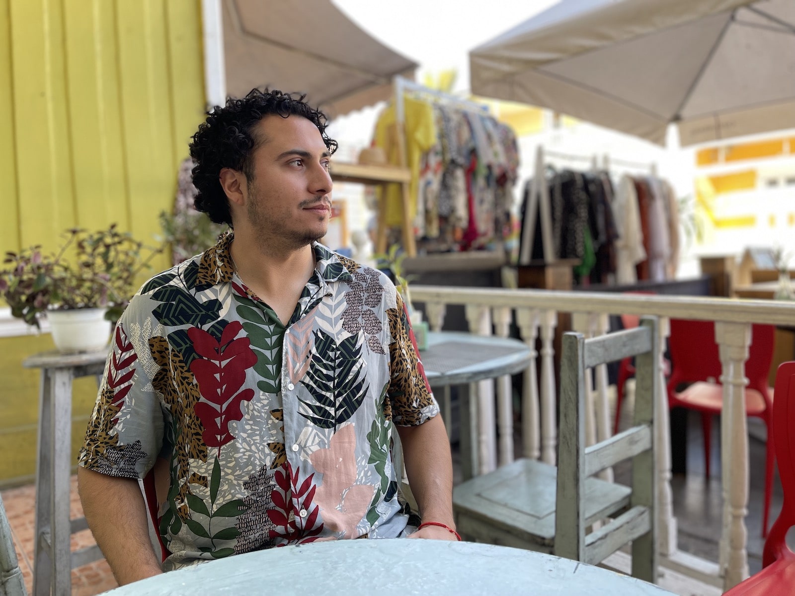 a man in a botannical-print shirt sits at a table near racks of clothes and outdoor umbrellas