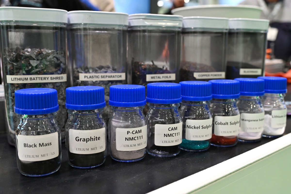 Small bottles labeled "black mass," "graphite," etc. are seen in a row in front of larger canisters with labels like "lithium battery shred"