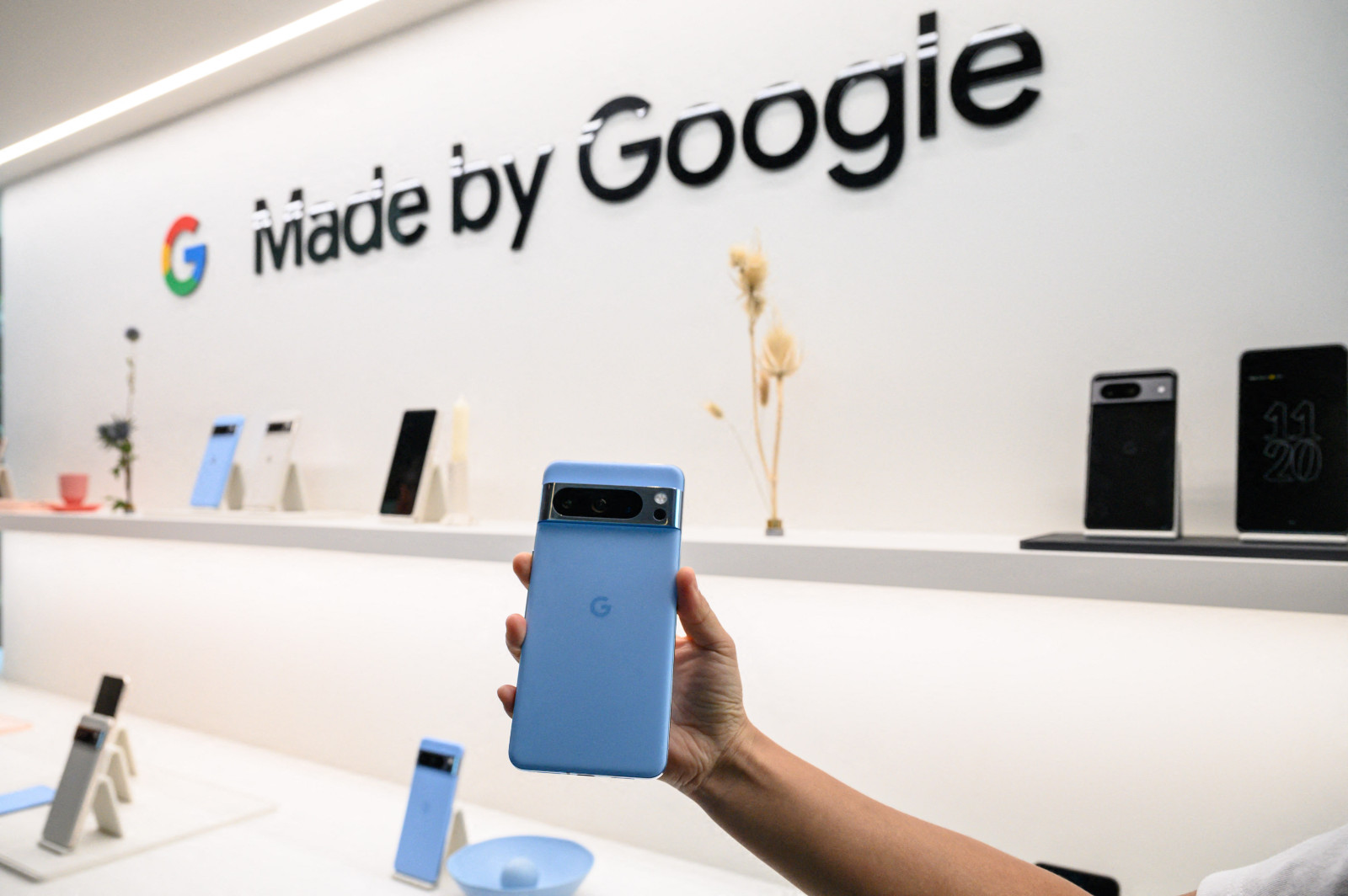 A hand holds a Pixel phone in a bright, white interior environment in front of a wall that says Made by Google