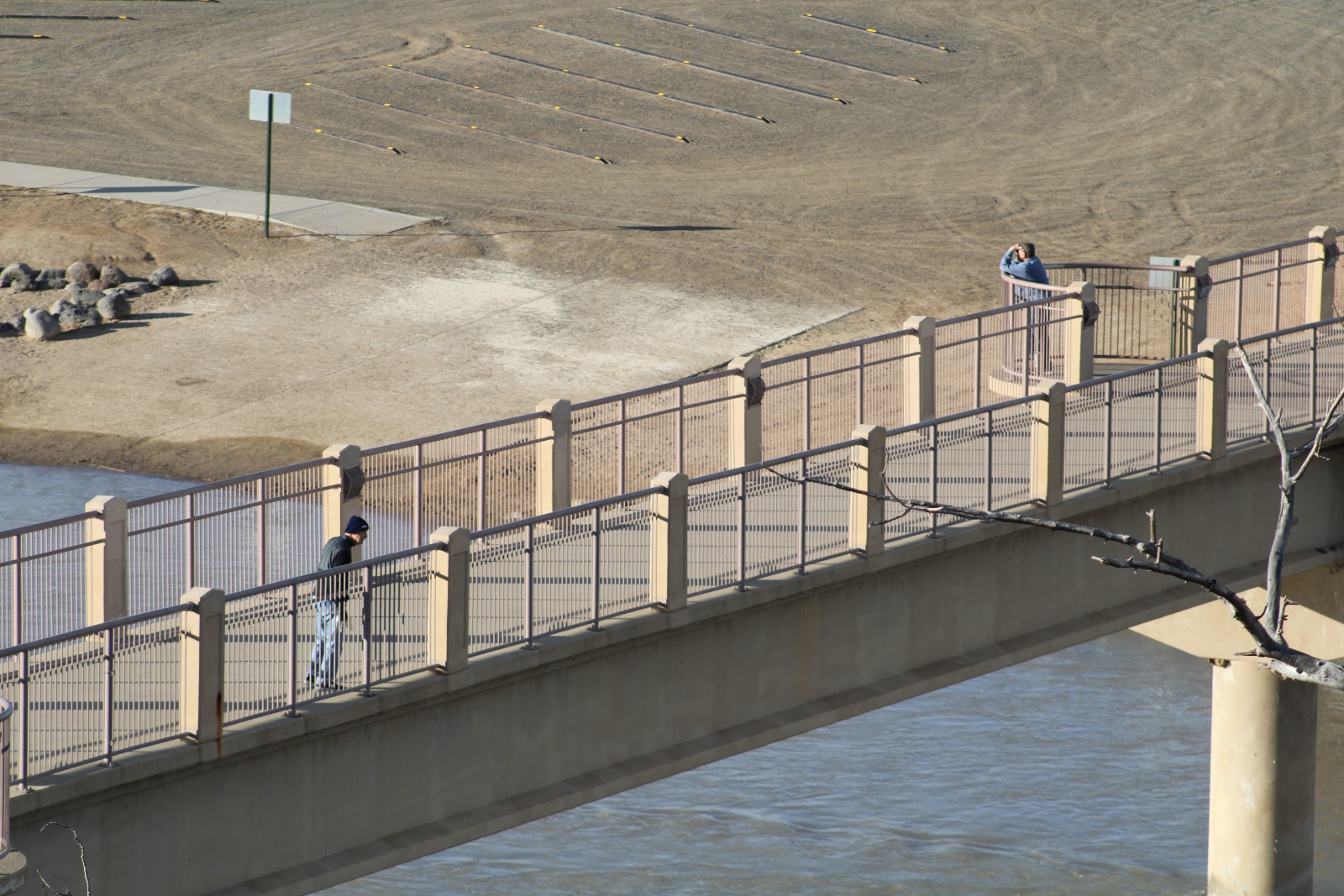 Two people walk over a narrow concrete brdige that overlooks sand and water.