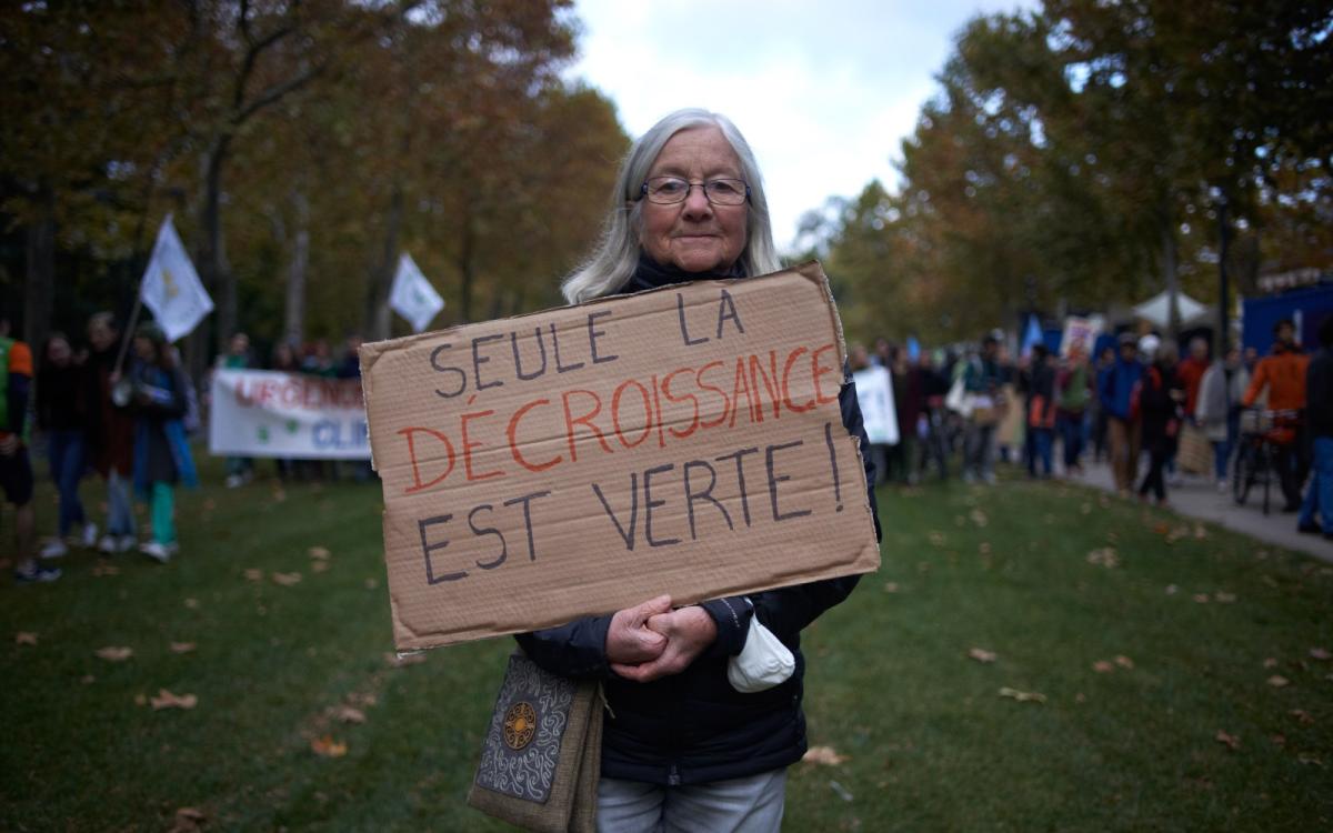 An older woman in a park faces the camera holding up a cardboard sign