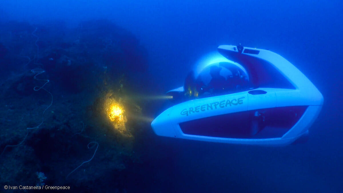 A submersible pod in dark blue waters shines a yellow light on a reef