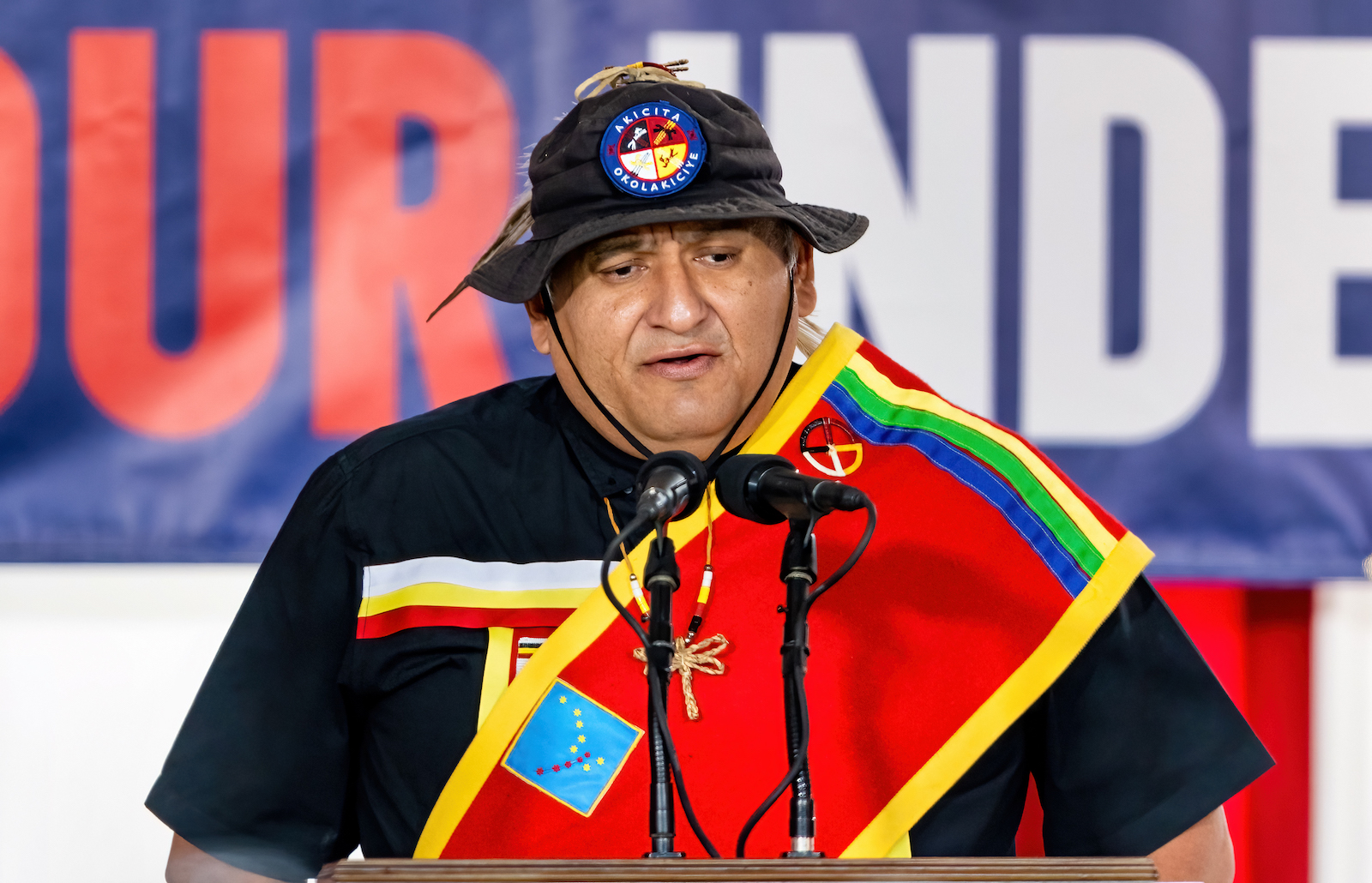 a man in a hat and colorful draped cloth stands in front of a microphone