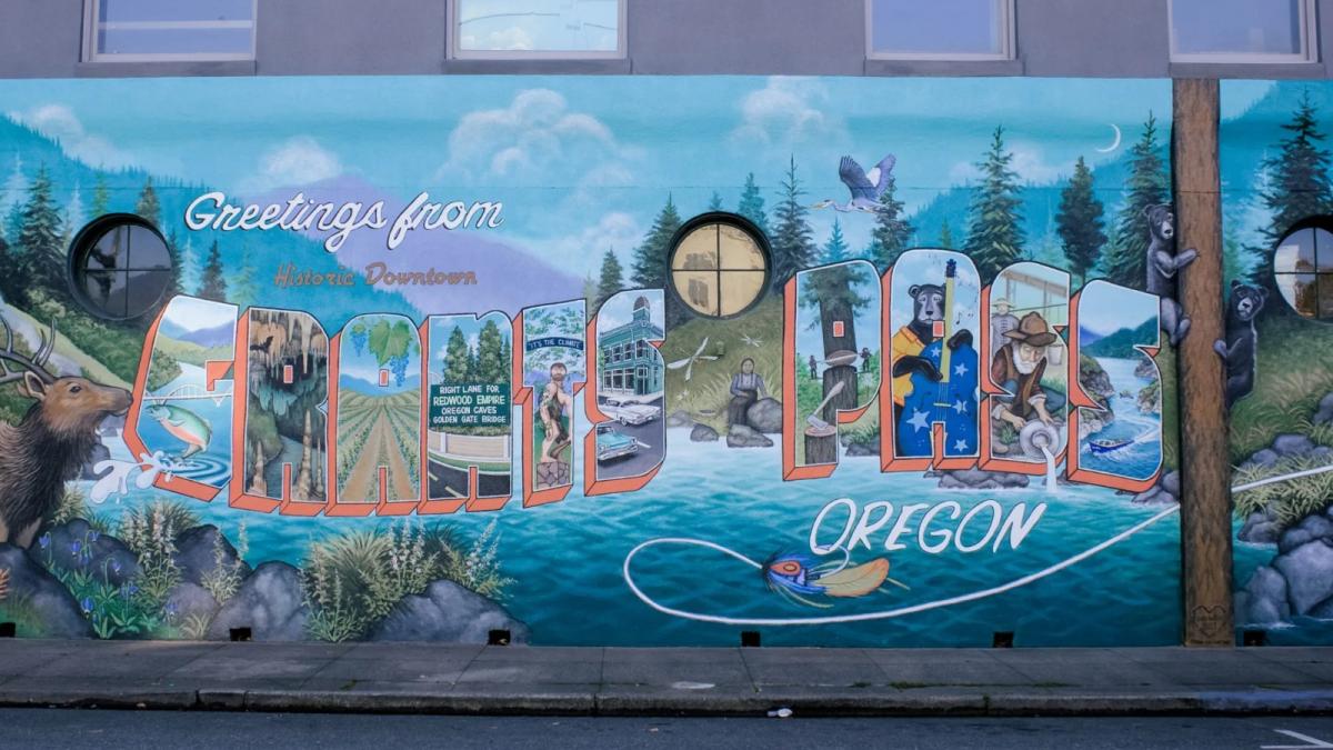 A mural that reads Greetings from Historic Downtown Grants Pass, Oregon, with a painting of a river and forest.