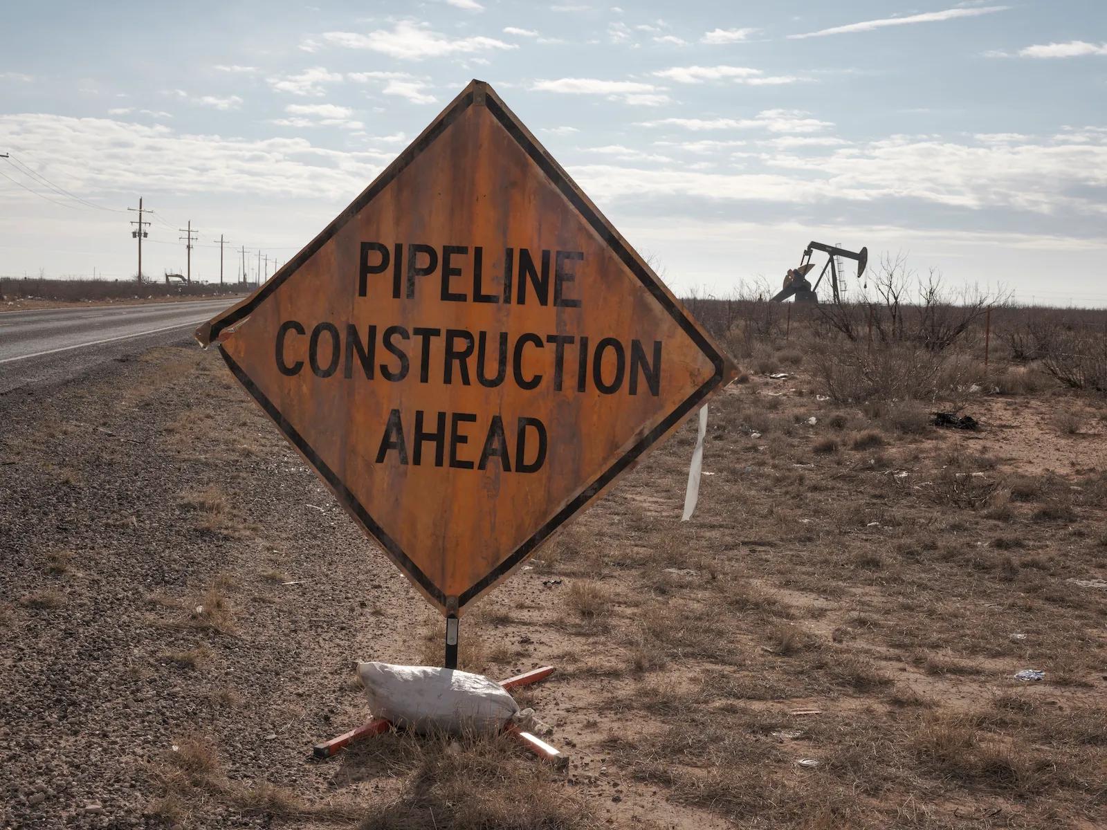 A diamond-shaped sign that says pipeline construction ahead with a pumpjack working in the distance