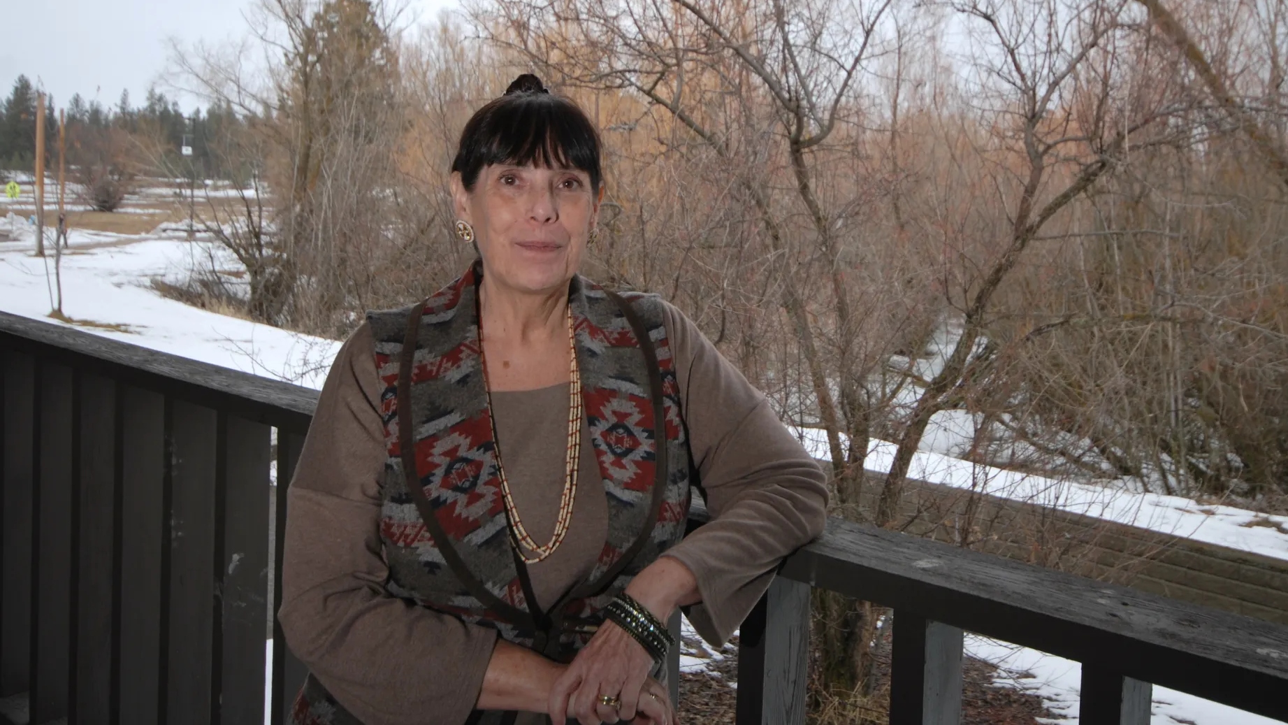 A woman with dark hair and bangs wears a brown shirt and a colorful scarf, and stands on a deck in front of a snowy yard.