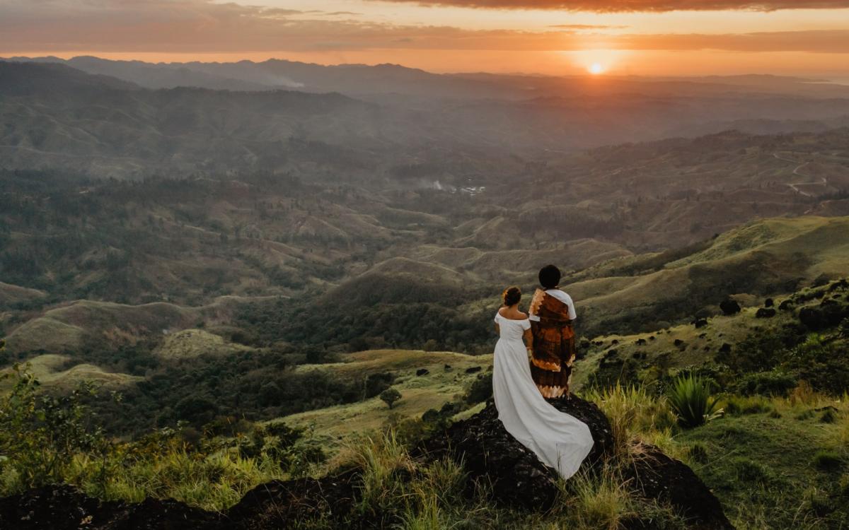 A bride and groom stand with their backs to the camera looking out at verdant green hills at sunset