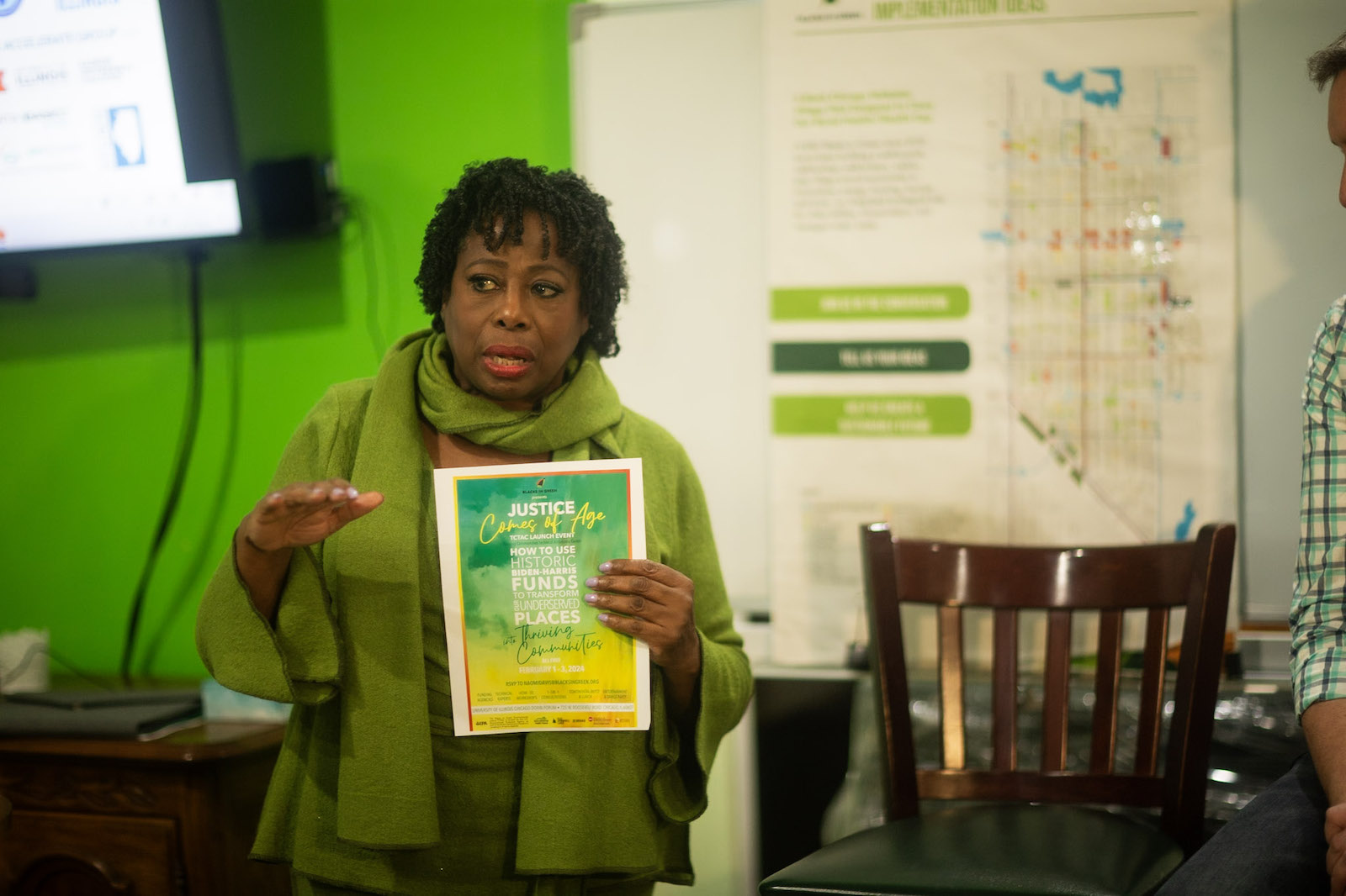 A woman in a green sweater holds a flyer while speaking in a bright green room