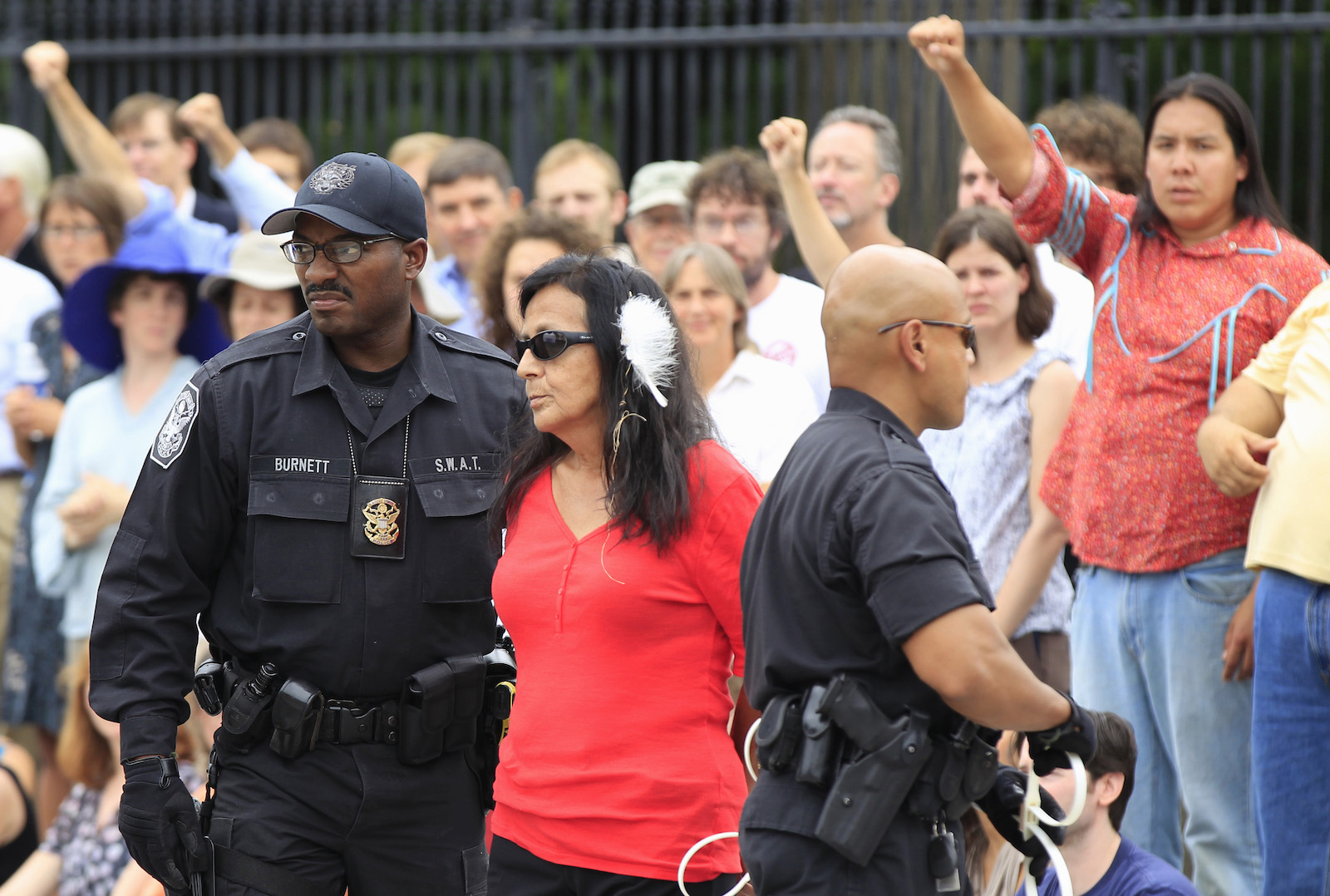 A woman in a red top and white feather in her hair is arrested in front of a crowd