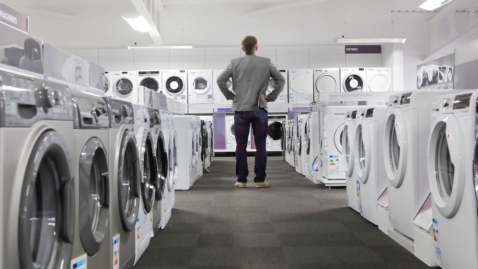 A man with his back to the camera considers a row of new washing machines and dryers in an appliance shop.