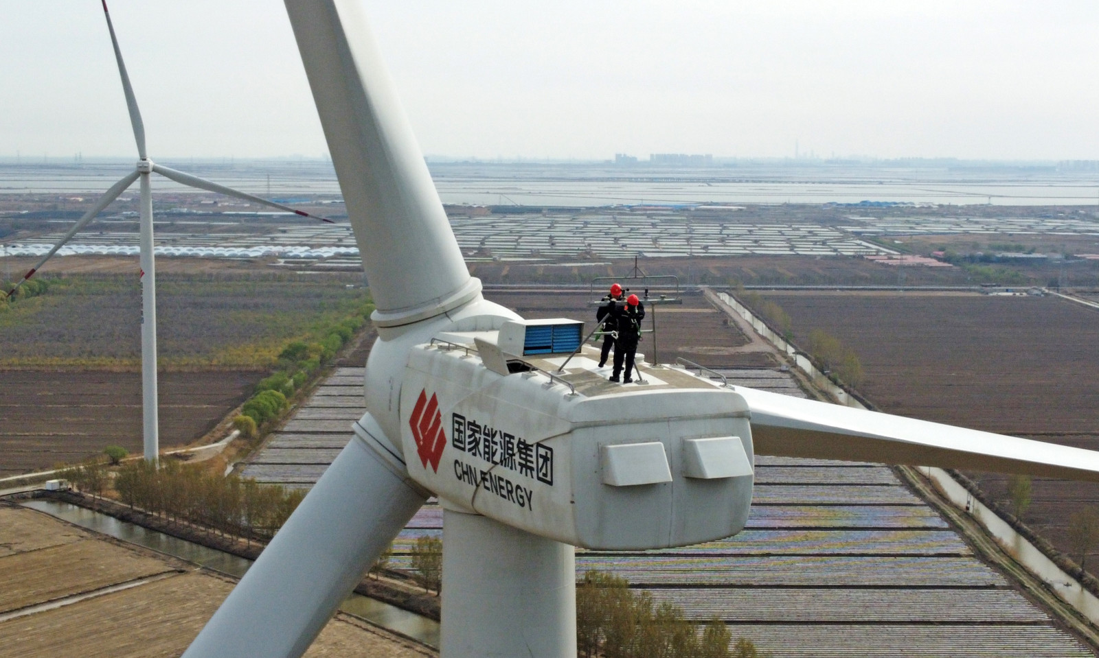 Two human figures are seen standing on a platform behind the three large blades of a wind turbine