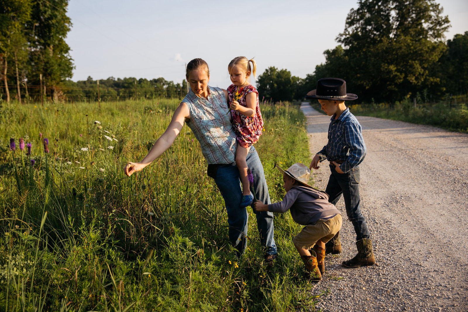 A woman holds a child near a field of tall grass and two children squat nearby