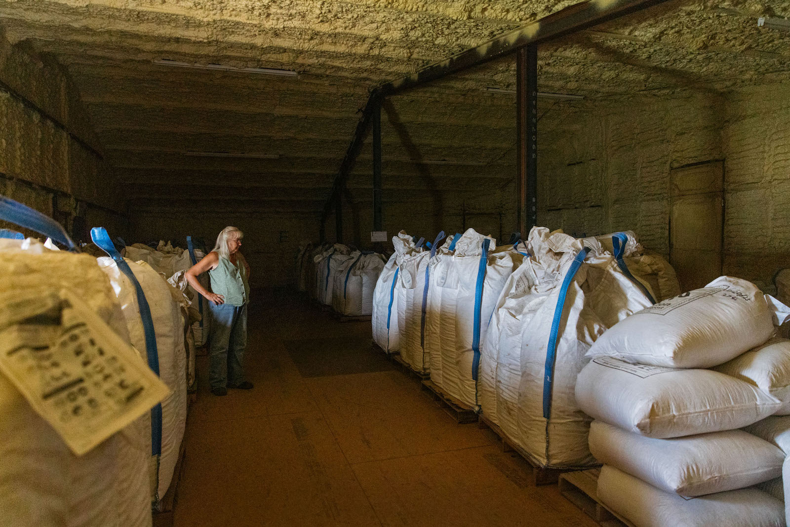 A senior woman stands in a large srorage room filled with stacks of large bags