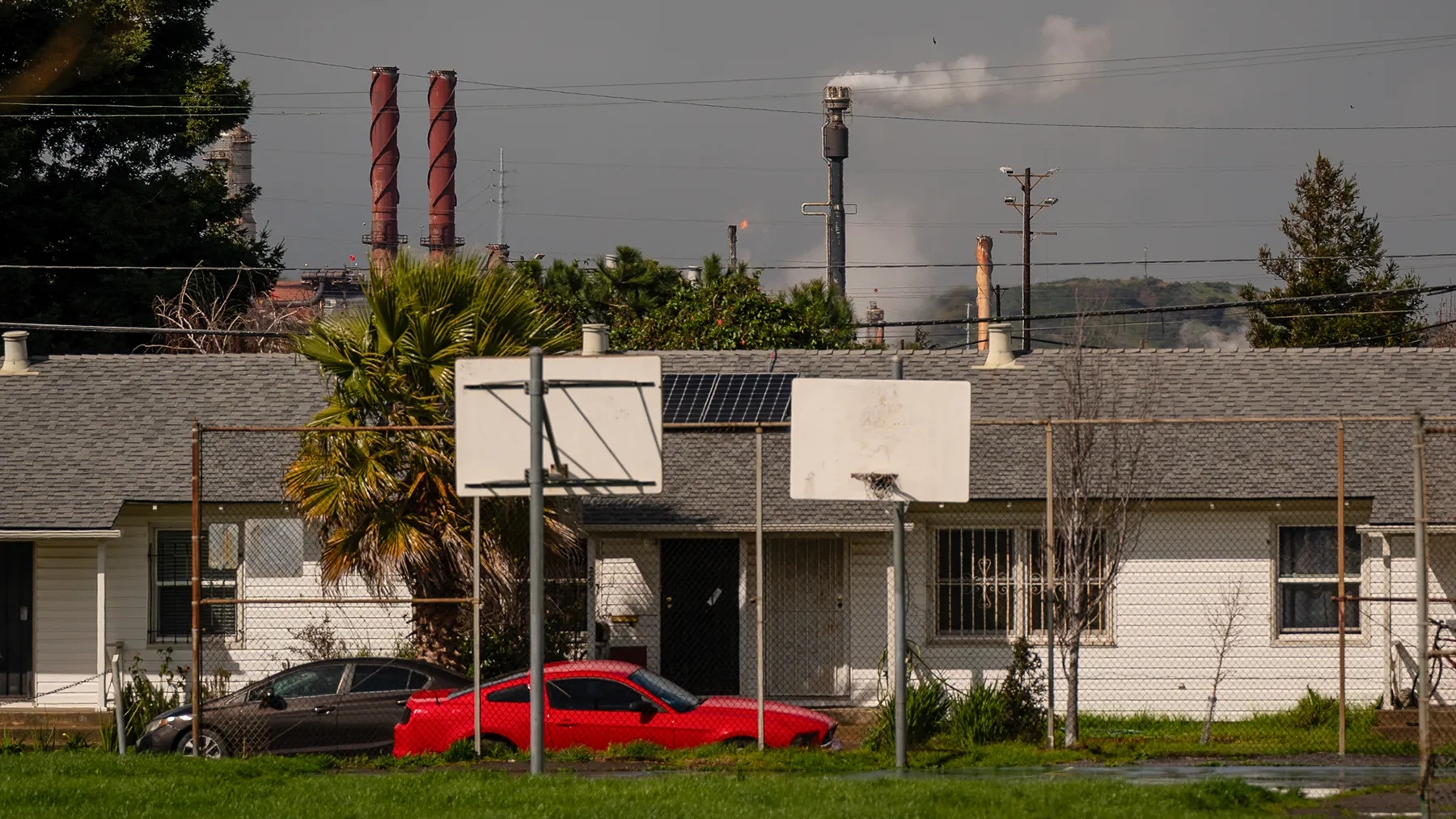 A small white house with a gray roof and solar panels sits in front of an industrial area with smokestacks. A red Mustang sits out front next to grass and basketball hoops.