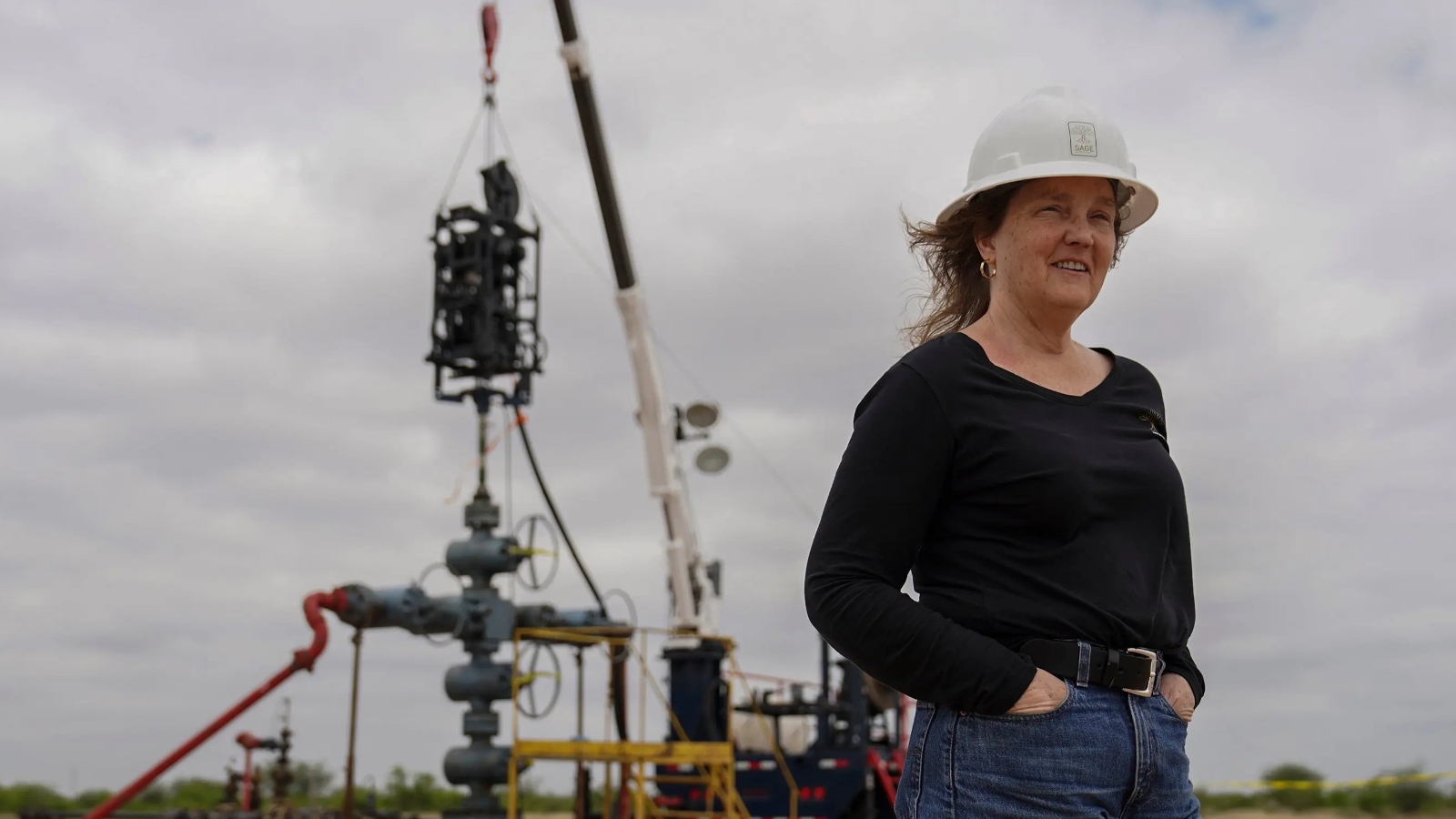 A woman in a long-sleeved black shirt and a white hard hat stands in front of an industrial construction site.
