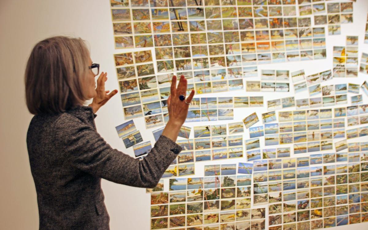 A woman gestures with her arms up, facing a wall where an array of vintage postcards are aligned and partially scattered