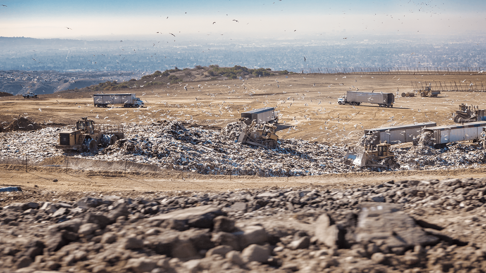 A landscape view of bulldozers and trucks working at a landfill while seagulls fly overhead