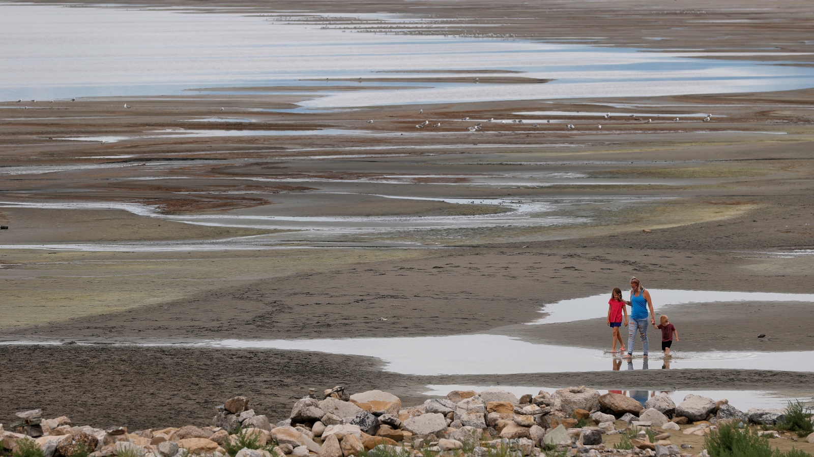 Three people walk on sand with puddles on the edge of a lake.