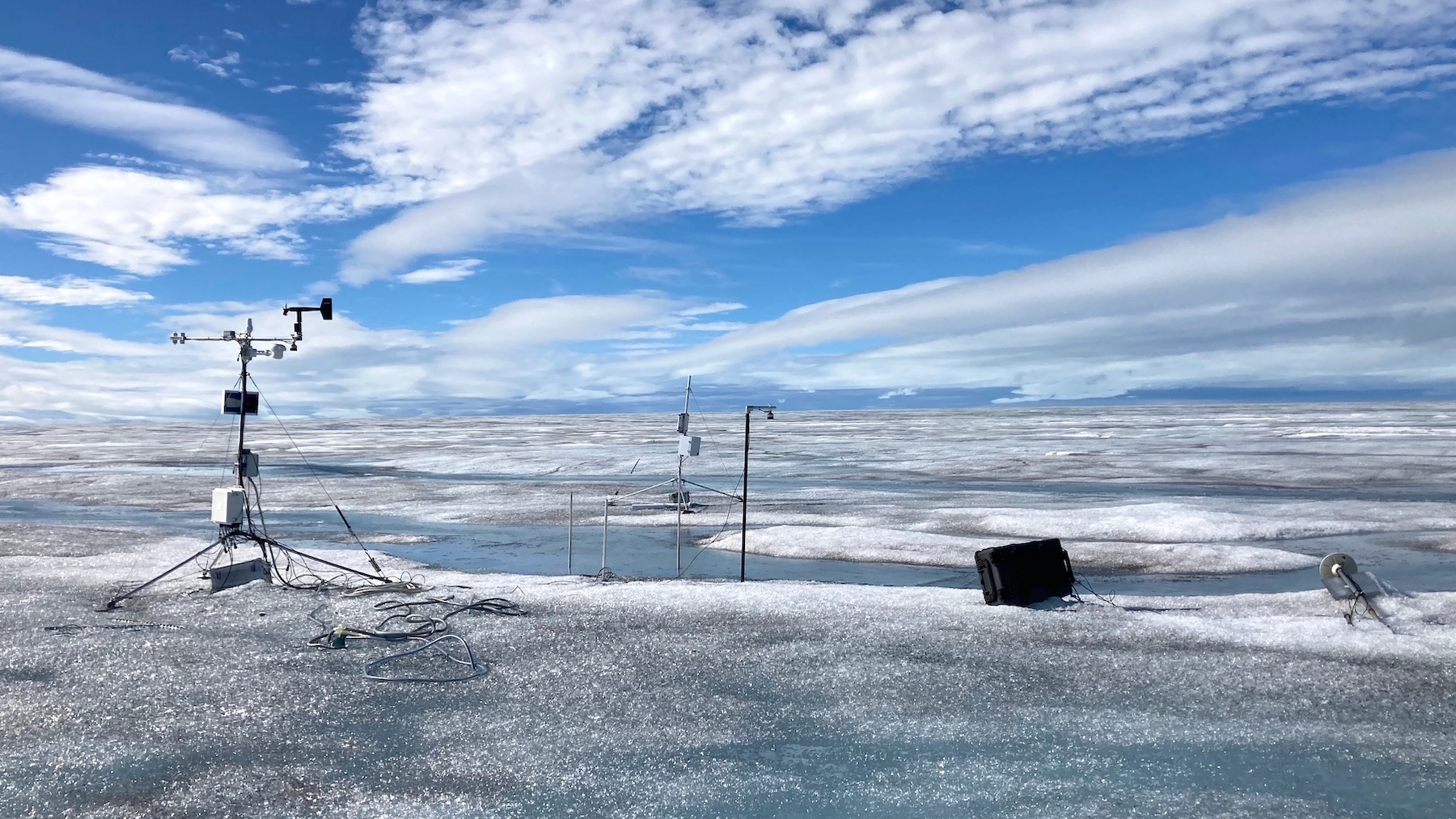 A sheet of ice stretches out in front of a blue sky. There is equipment sticking into the ice, and parts of it are melting.