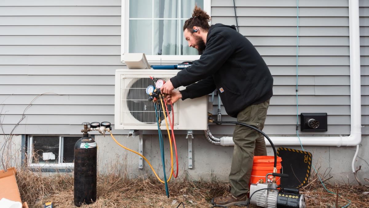 A man with dark hair in a bun, a black hoodie, and green pants installs a piece of equipment outside a house.