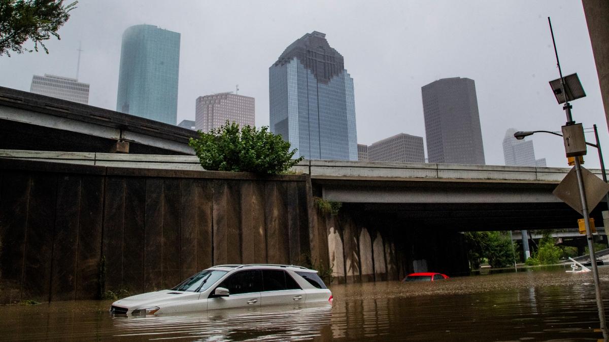 A silver SUV sits in several feet of water below a highway and a city skyline.