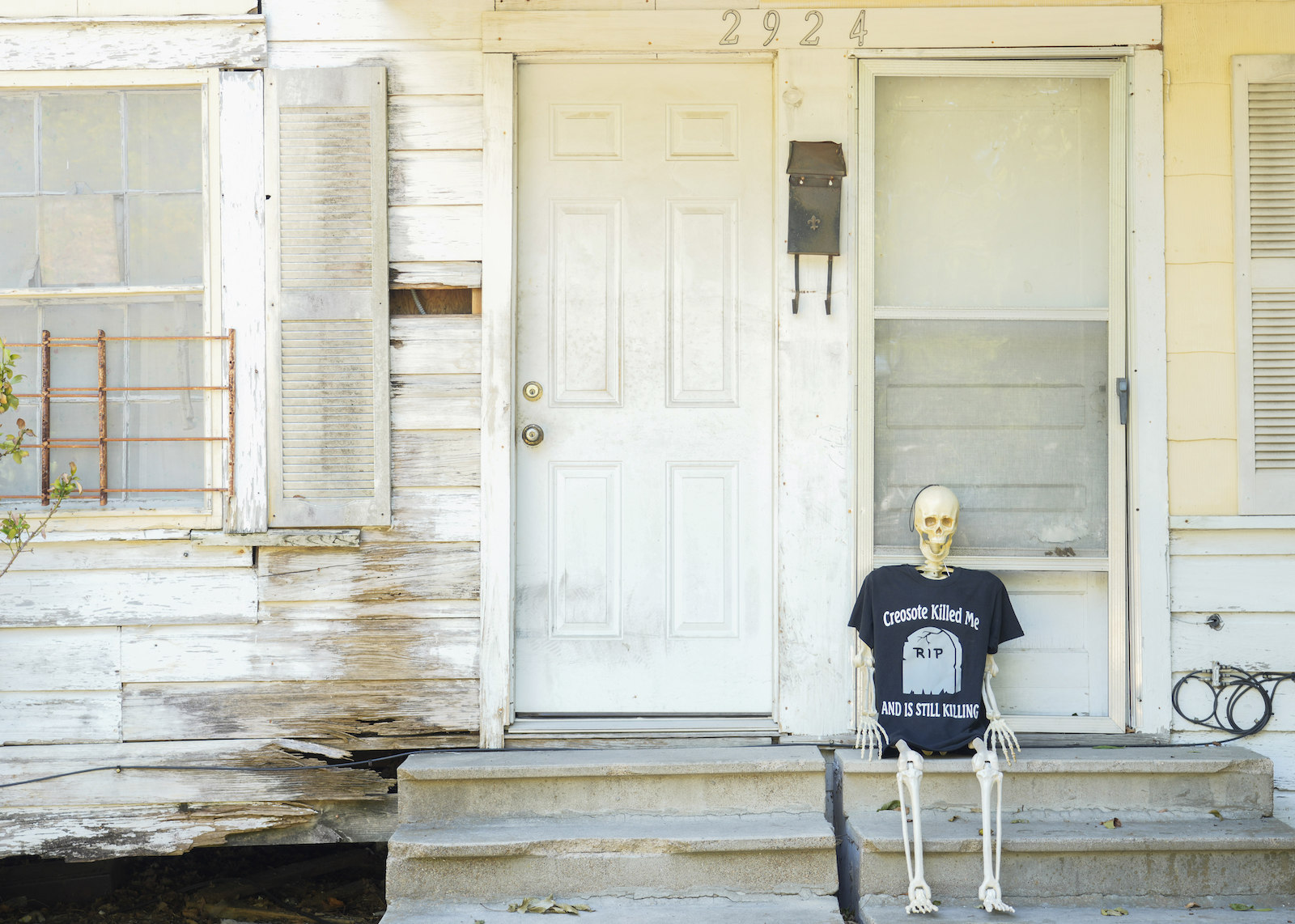 A plastic skeleton wearing a black t-shirt with "creosote killed me" on it sits on a porch