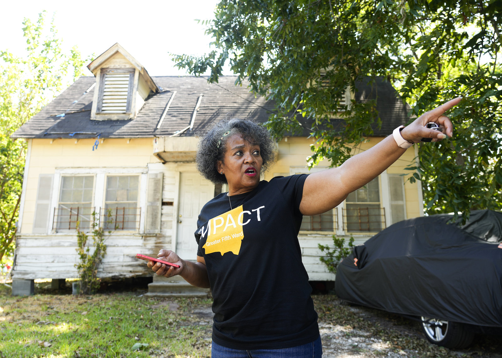 A woman in a black t-shirt with a yellow logo points beyond the camera while standing in front of a house