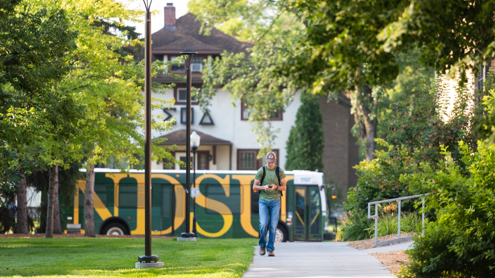 A young man in jeans and a tee shirt walks on a path in front of a frat house and a bus that reads NDSU.