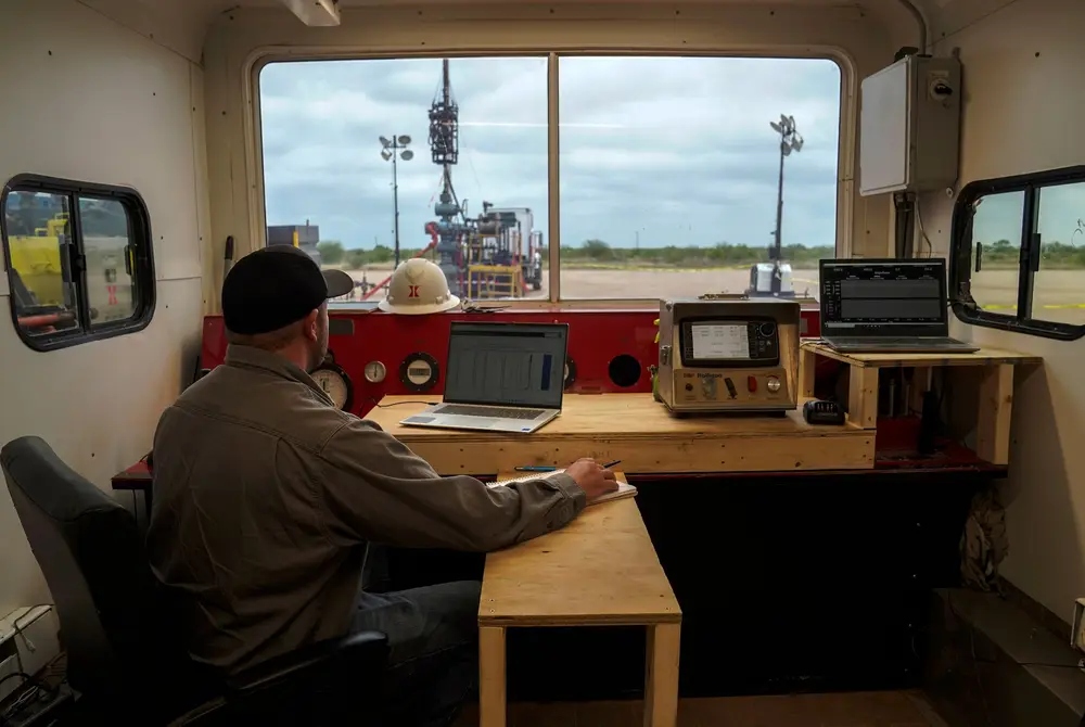 A man sits at a desk with a large window looking out over an industrial site.