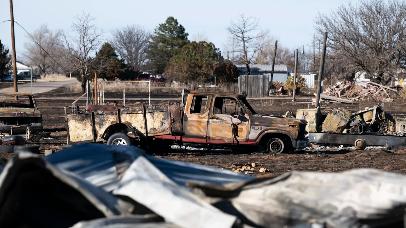 A charred pickup truck sits in front of a pile of debris amid a scorched landscape.