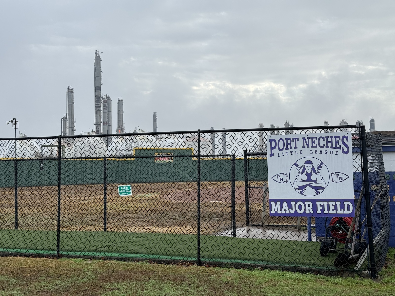 Sign reading "Port Neches Little League Major Field" in foreground with petrochemical complex in background