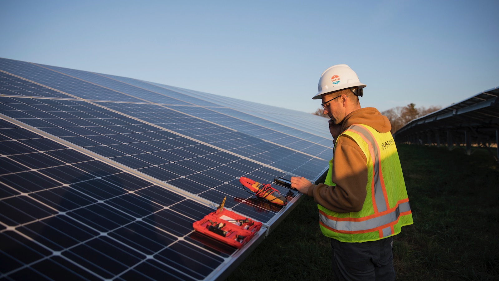 An electrician in a yellow vest and whote hard hat works on a photovoltaic panel in a solar array on a farm in Massachusetts.