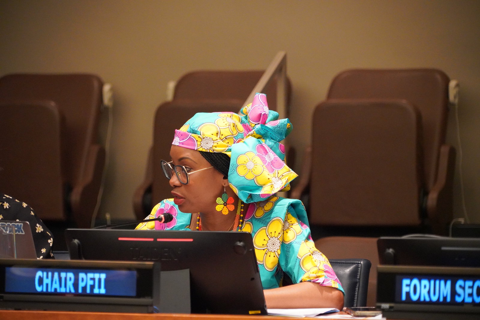 A woman wears a matching floral shirt and sheet headdress and sits at a desk with a label that says Chair PFII