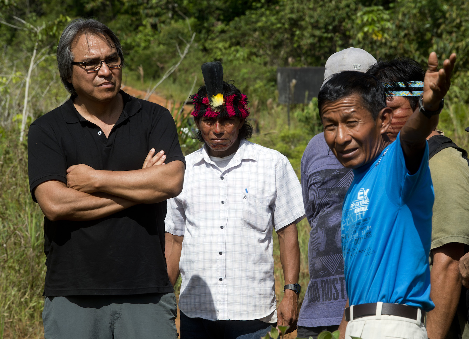 A man in a black shirt crosses his arms while talking to a group of other people. One person in a blue shirt gestures beyond the scene to the surrounding wilderness