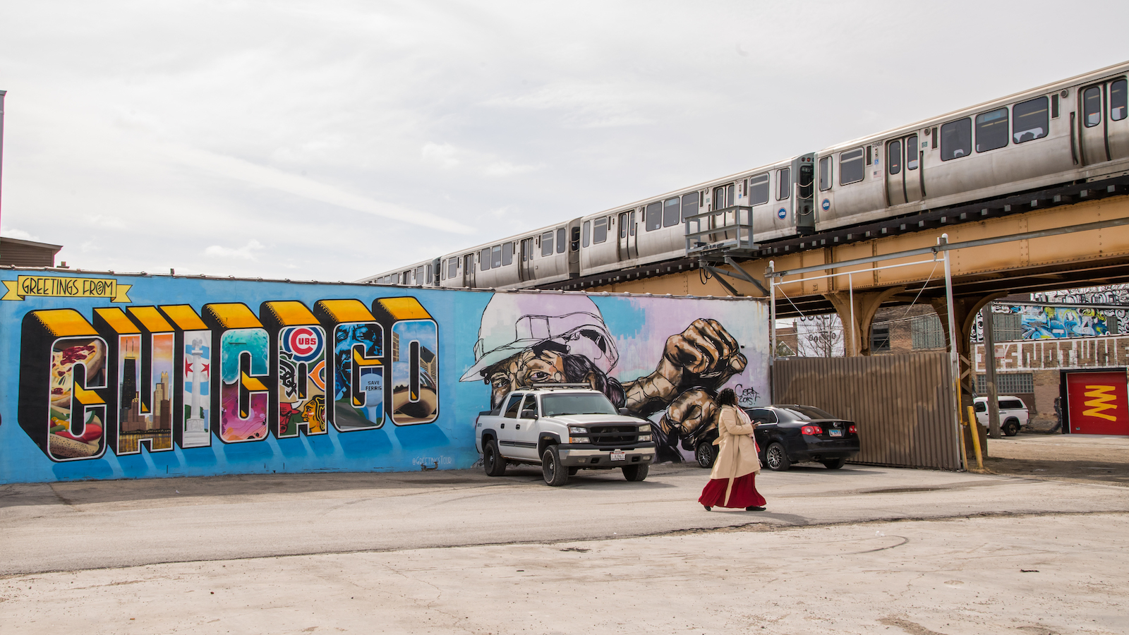 A mural with the world Chicago on it next to an elevated train track and woman walking across a parking lot.