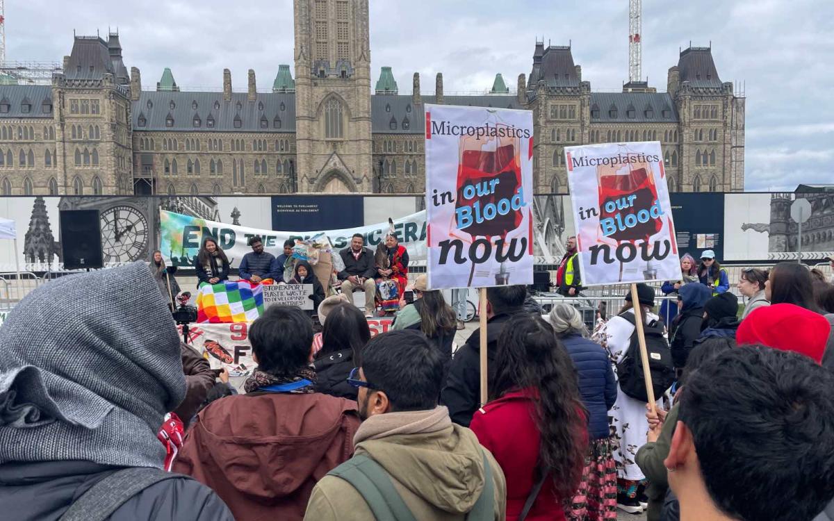 In the foreground, a crowd stands before a stage where a handful of leaders are sitting, waiting to speak. In the background is Canada's Parliament Hill.