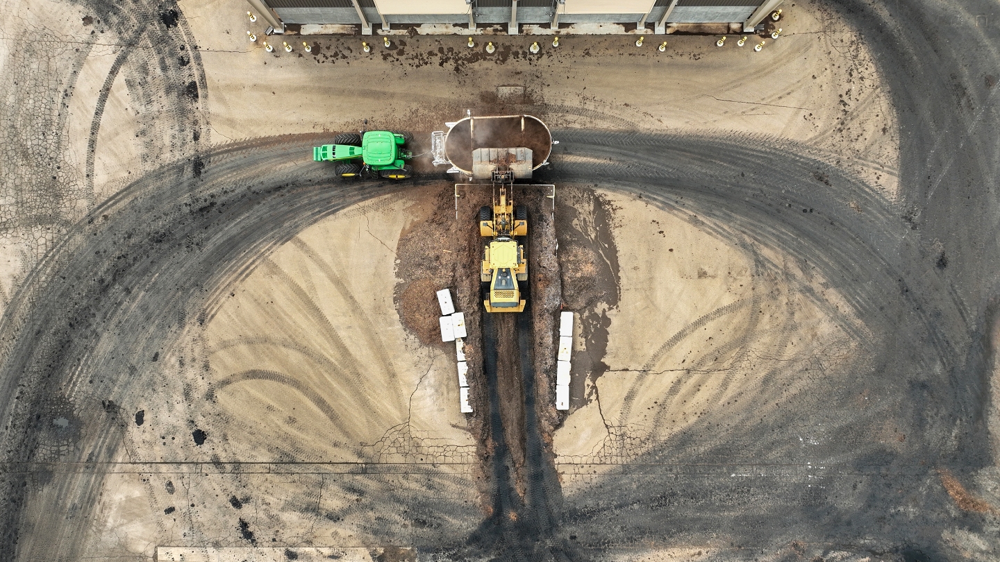 An aerial shot of an outdoor industrial with green and yellow construction equipment next to a vat containing brown material