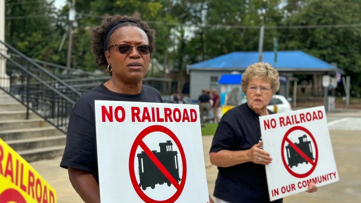 A Black woman with short hair and sunglasses stands next to a white woman with short blonde hair and glasses. They both hold signs that say No Railroad in our community.